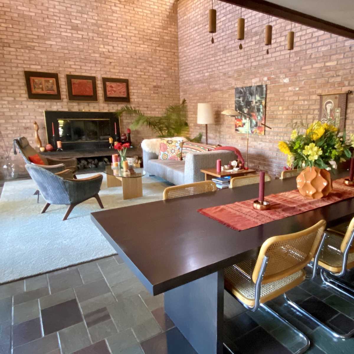 A modern living room features a brick wall, a fireplace, a dining table with chairs, and various decorative elements.