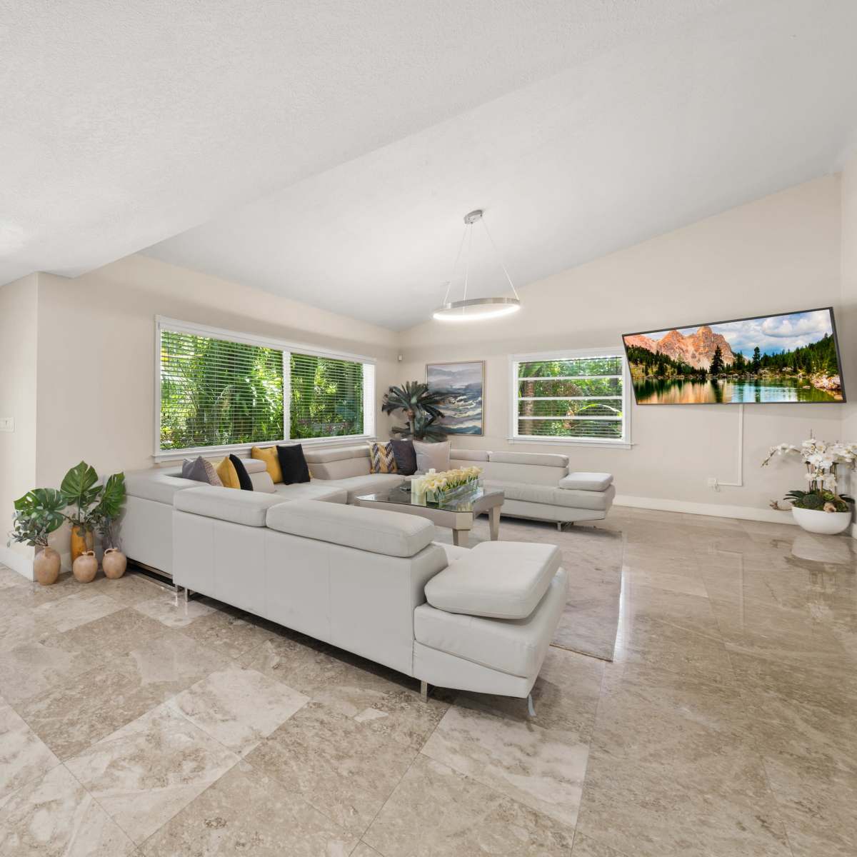 A bright living room features a sectional sofa, a wall-mounted TV, and large windows overlooking a garden.