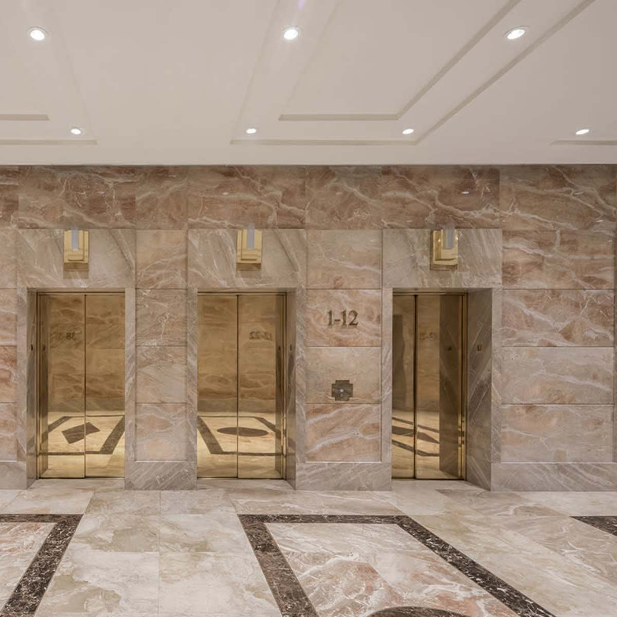 A spacious lobby with three elevator doors set in a wall of beige stone and marble flooring.