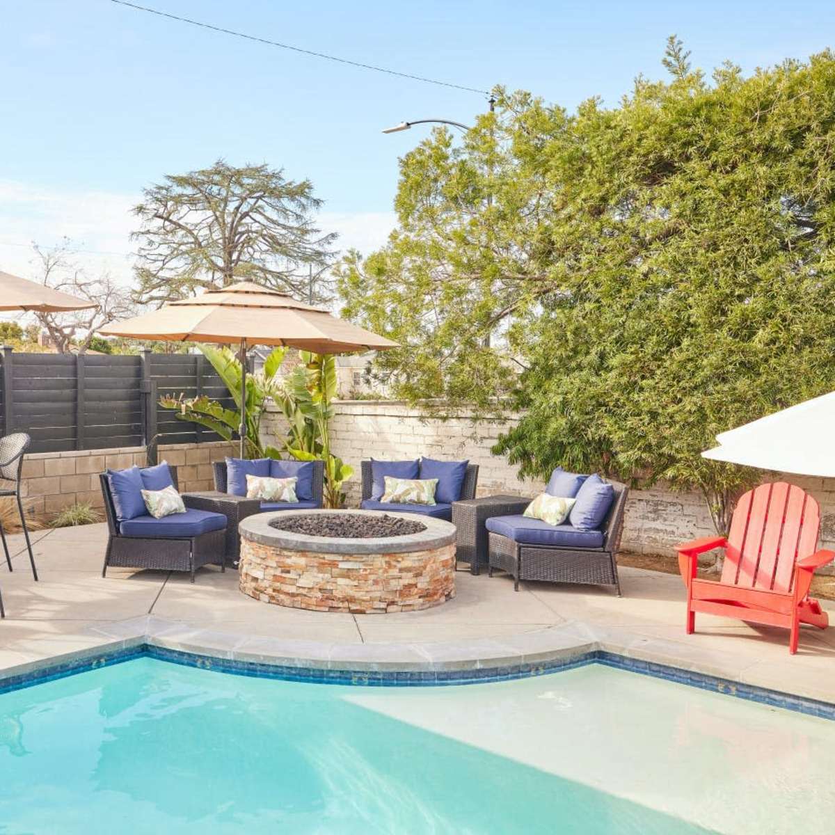 A backyard with a swimming pool, a stone fire pit surrounded by seating, and patio umbrellas providing shade.