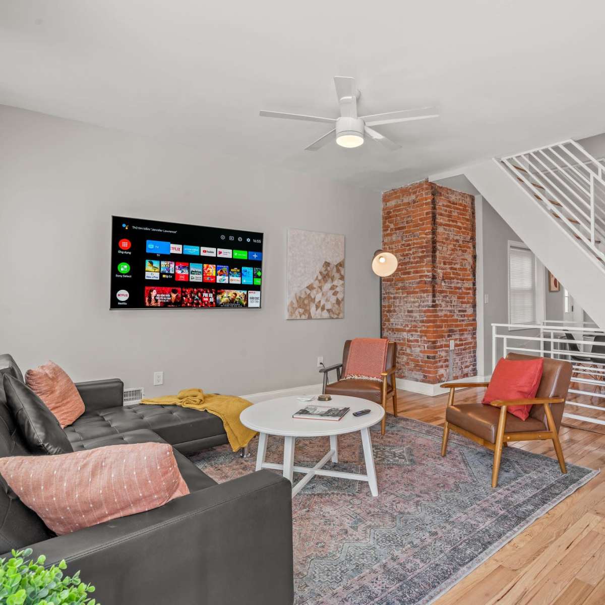 The living room features a modern design with a large wall-mounted TV, a sectional sofa, two accent chairs, a round coffee table, and a staircase leading to the upper level, complemented by exposed brick accents.