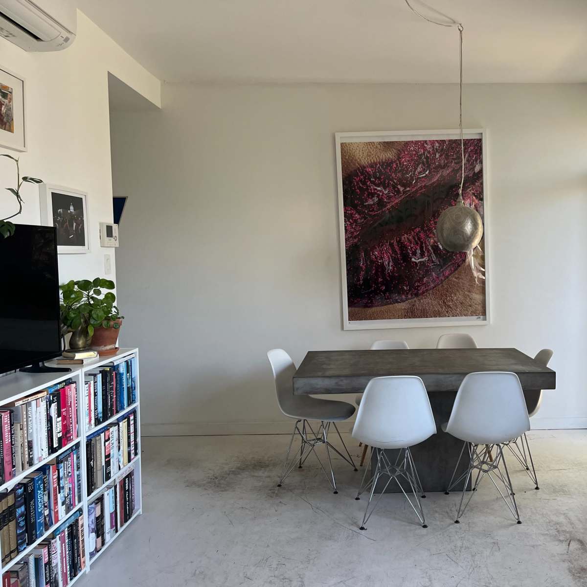 A minimalist dining area with a large art piece on the wall, a concrete dining table surrounded by white chairs, and a bookshelf filled with books.