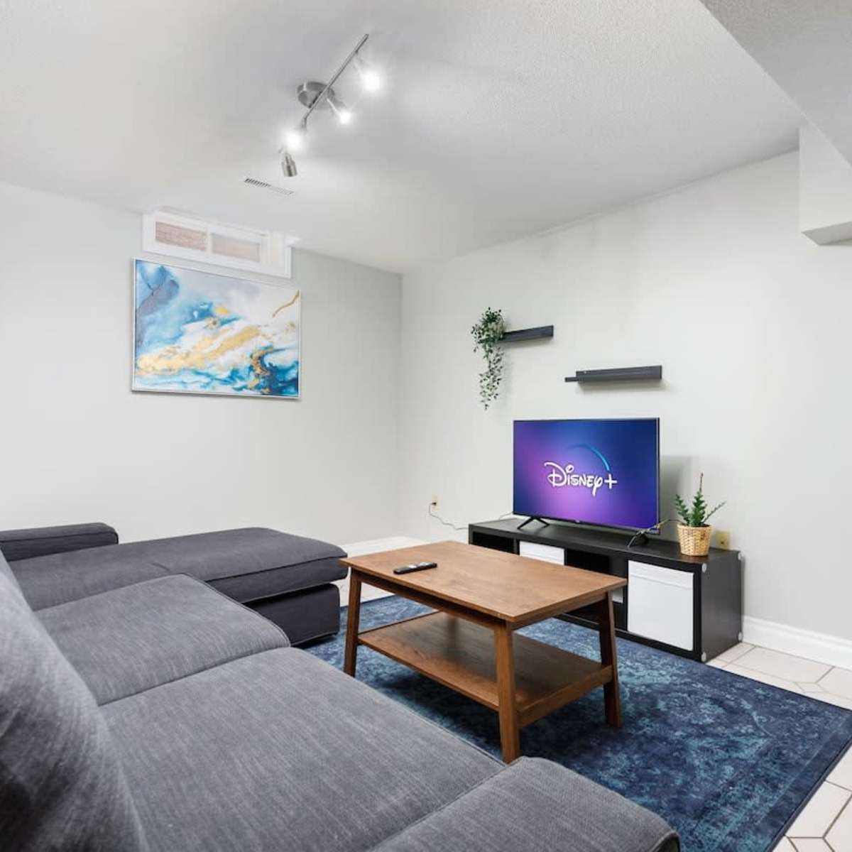 A modern living room with a dark gray sectional sofa, a wooden coffee table, and a TV stand featuring a screen displaying the Disney+ logo.