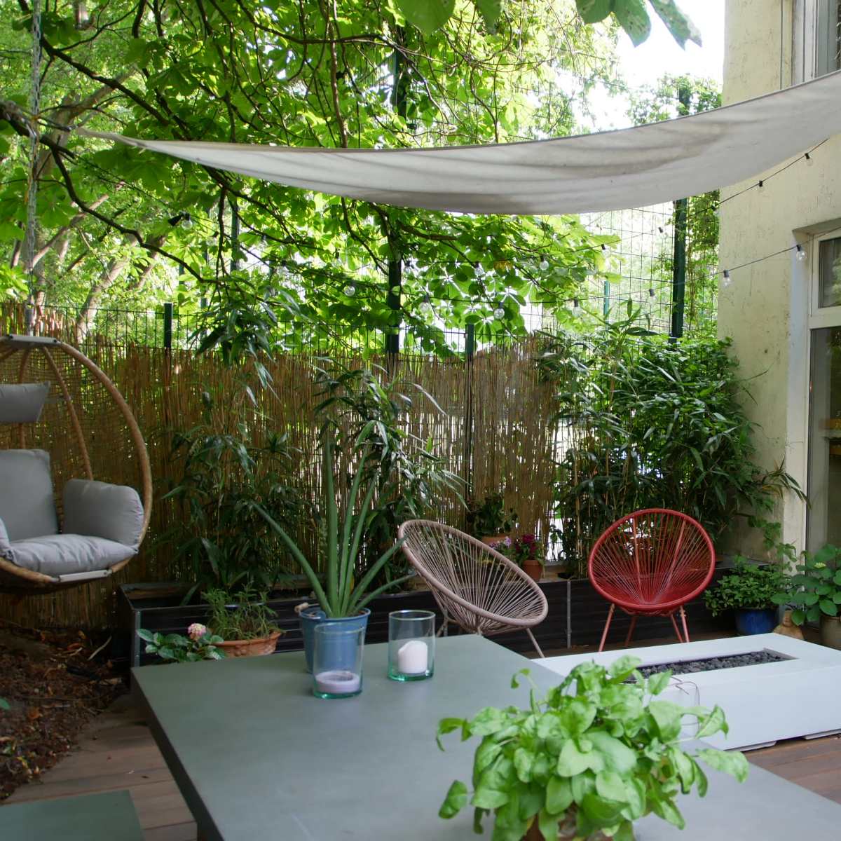 A patio area features a shaded table, potted plants, and colorful chairs surrounded by greenery and a house.