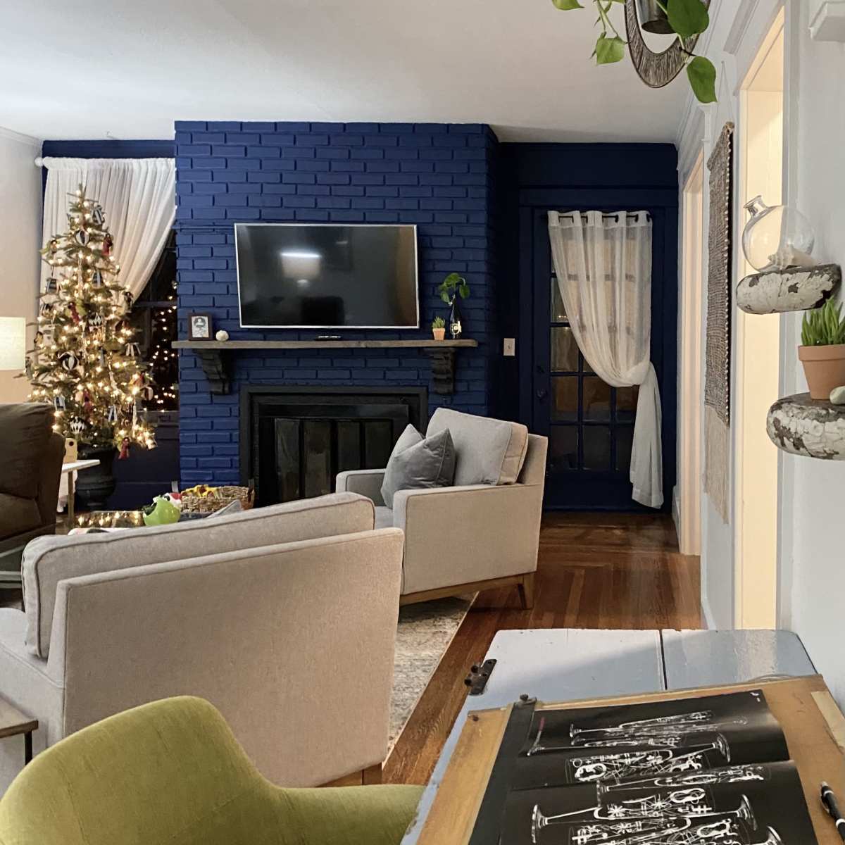 A cozy living room features a dark blue brick wall, a decorated Christmas tree, and a writing desk with sheet music in the foreground.