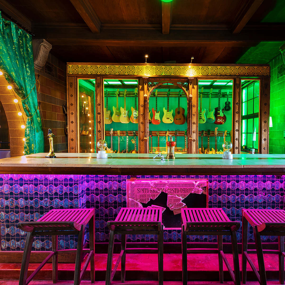 A colorful bar area featuring a wooden counter, several bar stools, and a display of guitars in a vibrant setting with green and purple lighting.