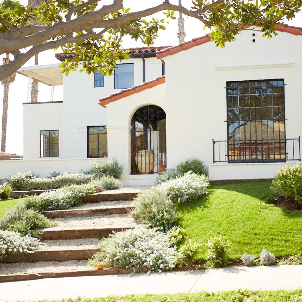 A two-story white house with a red-tiled roof is situated on a grassy lawn, featuring a stone walkway leading to the entrance.