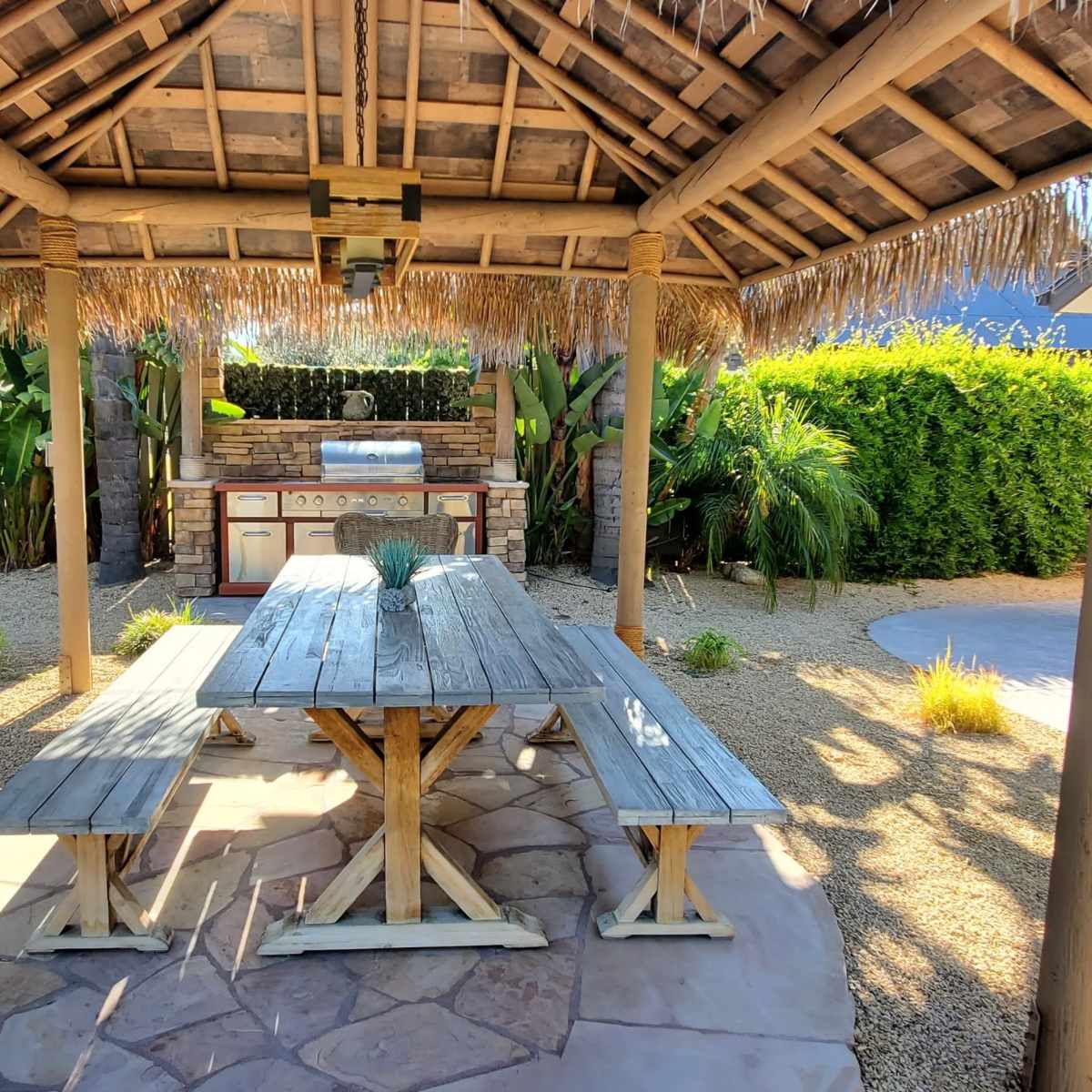 An outdoor patio with a thatched roof, a wooden picnic table, and an outdoor kitchen area surrounded by landscaping.