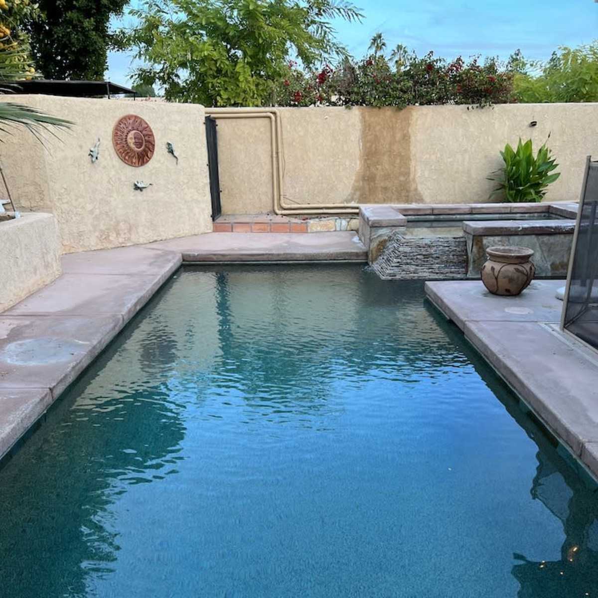 A clear swimming pool surrounded by stone patios, with a fence and lush greenery in the background.
