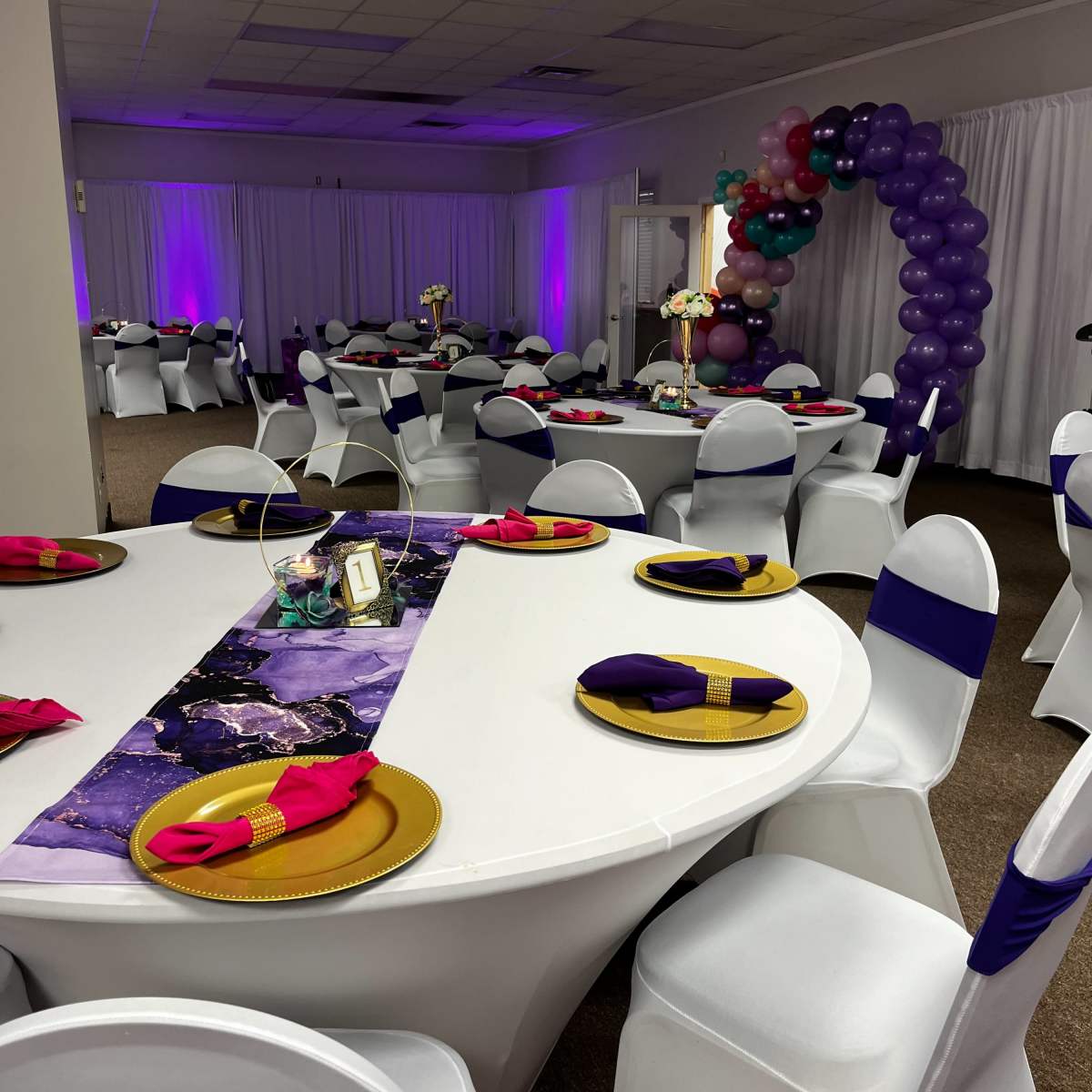 A decorated event space with tables arranged for a gathering, featuring white tablecloths, colorful napkins, and a balloon arch in the background.