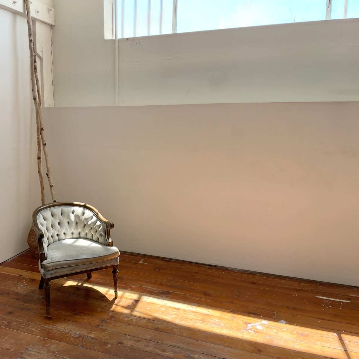 A vintage upholstered chair is positioned in a sunlit corner of a minimalist room with wooden flooring and white walls.