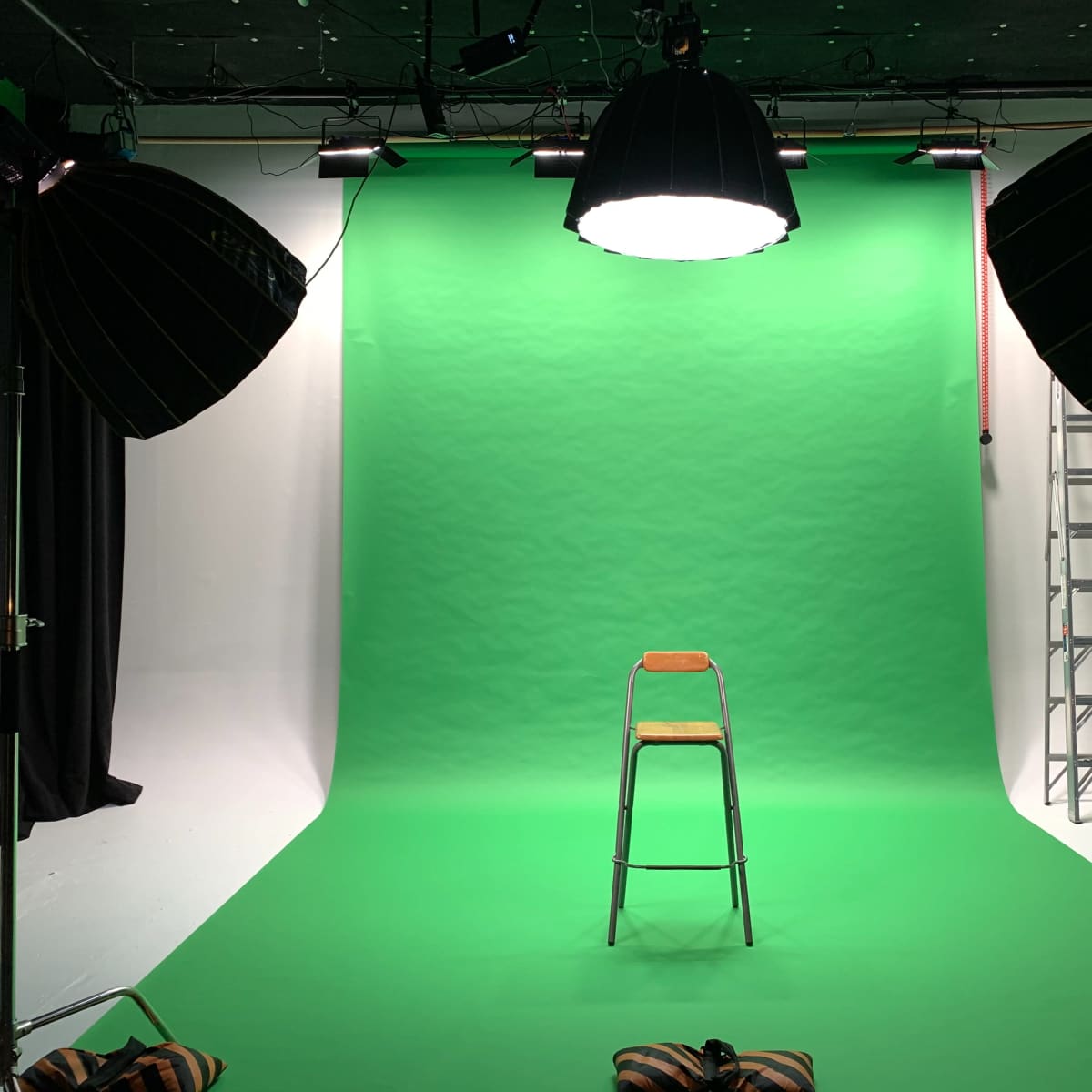 A studio setup features a green screen backdrop with a metal chair in the center, flanked by large studio lights and a ladder in the background.