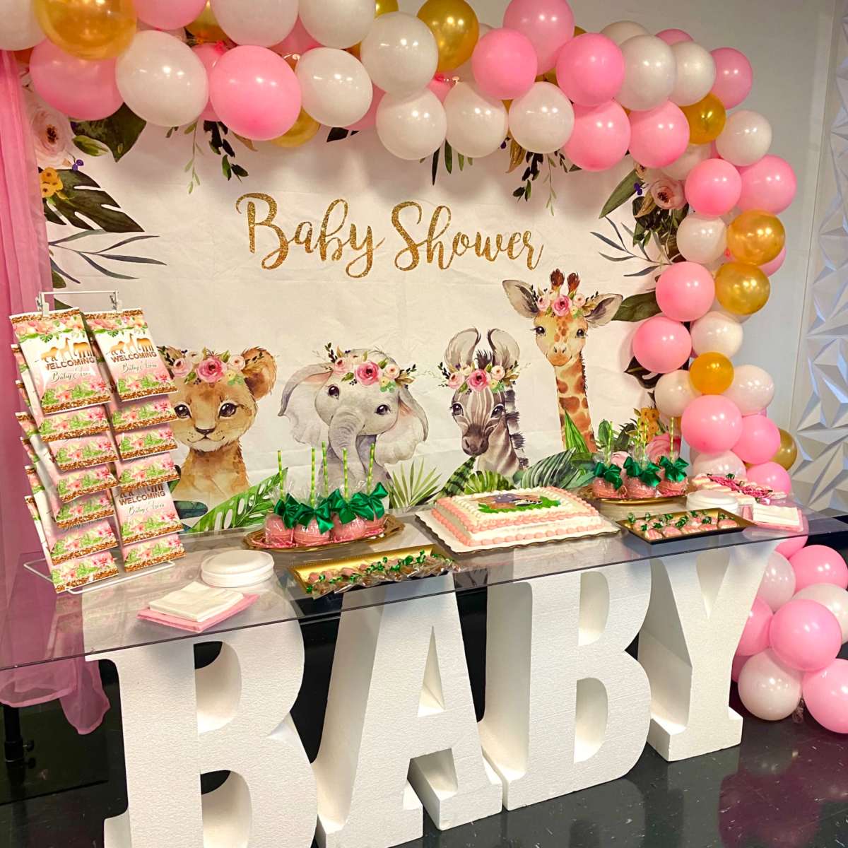 A decorated table for a baby shower featuring a cake, treats, and a backdrop with animal illustrations and balloons in pink and gold.