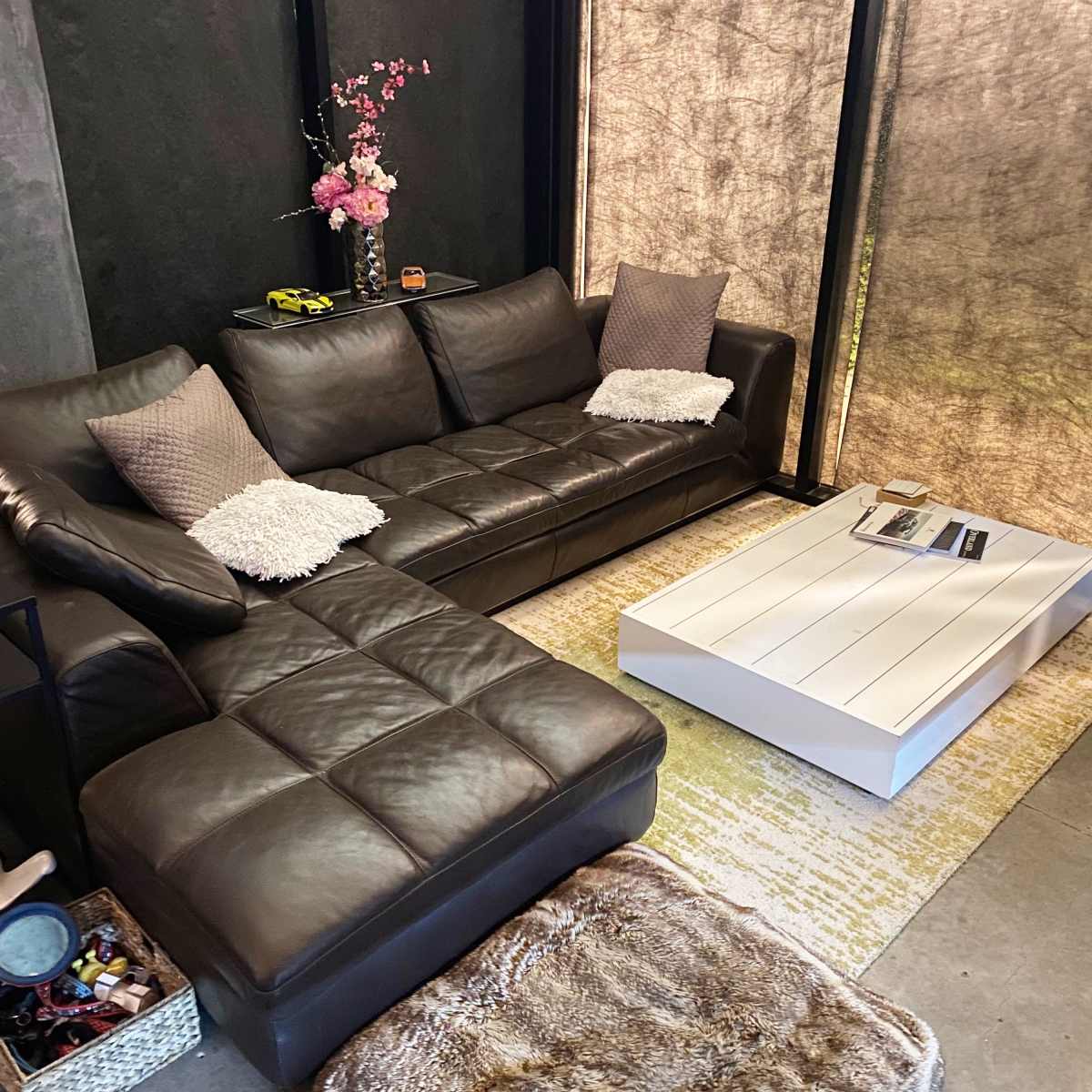 A modern living room featuring a dark leather sectional sofa, a white coffee table, and decorative accents on a gray wall with large windows covered by sheer curtains.