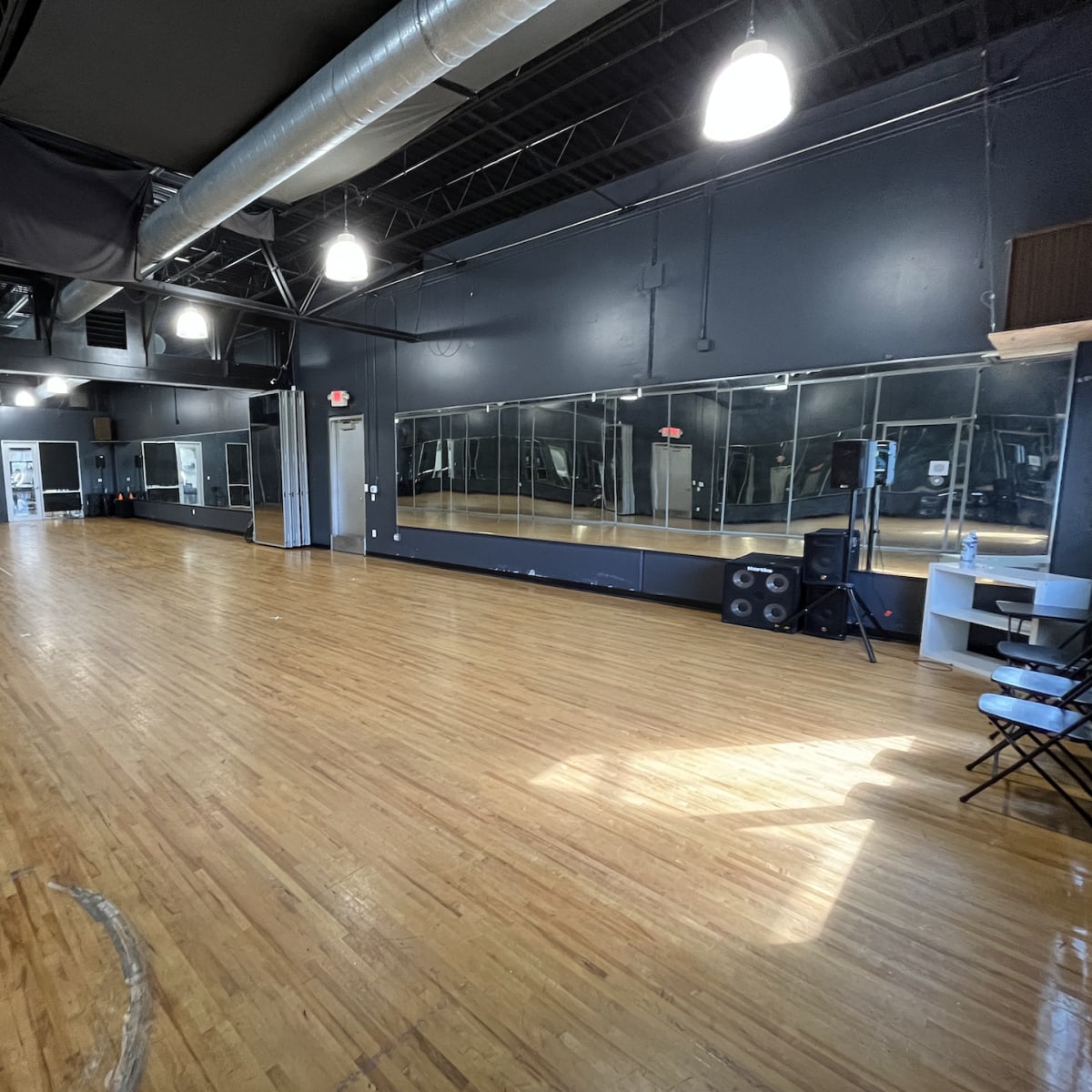 A spacious dance studio with a wooden floor, large mirrors on one wall, and a sound system setup in one corner.