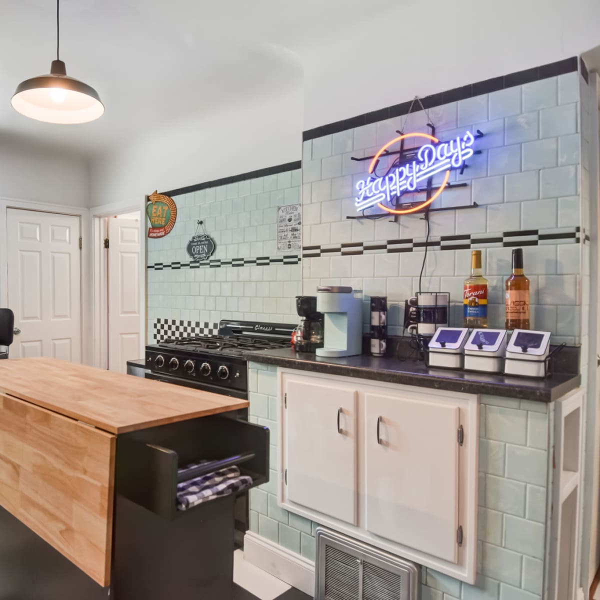The kitchen features a checkered black-and-white floor, a wooden island, and retro-style decor with blue tiles and a neon sign.