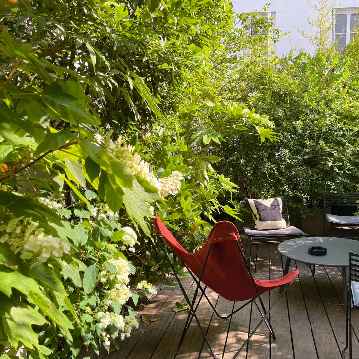 A small, lush green outdoor seating area with a round table and a red chair surrounded by dense foliage.