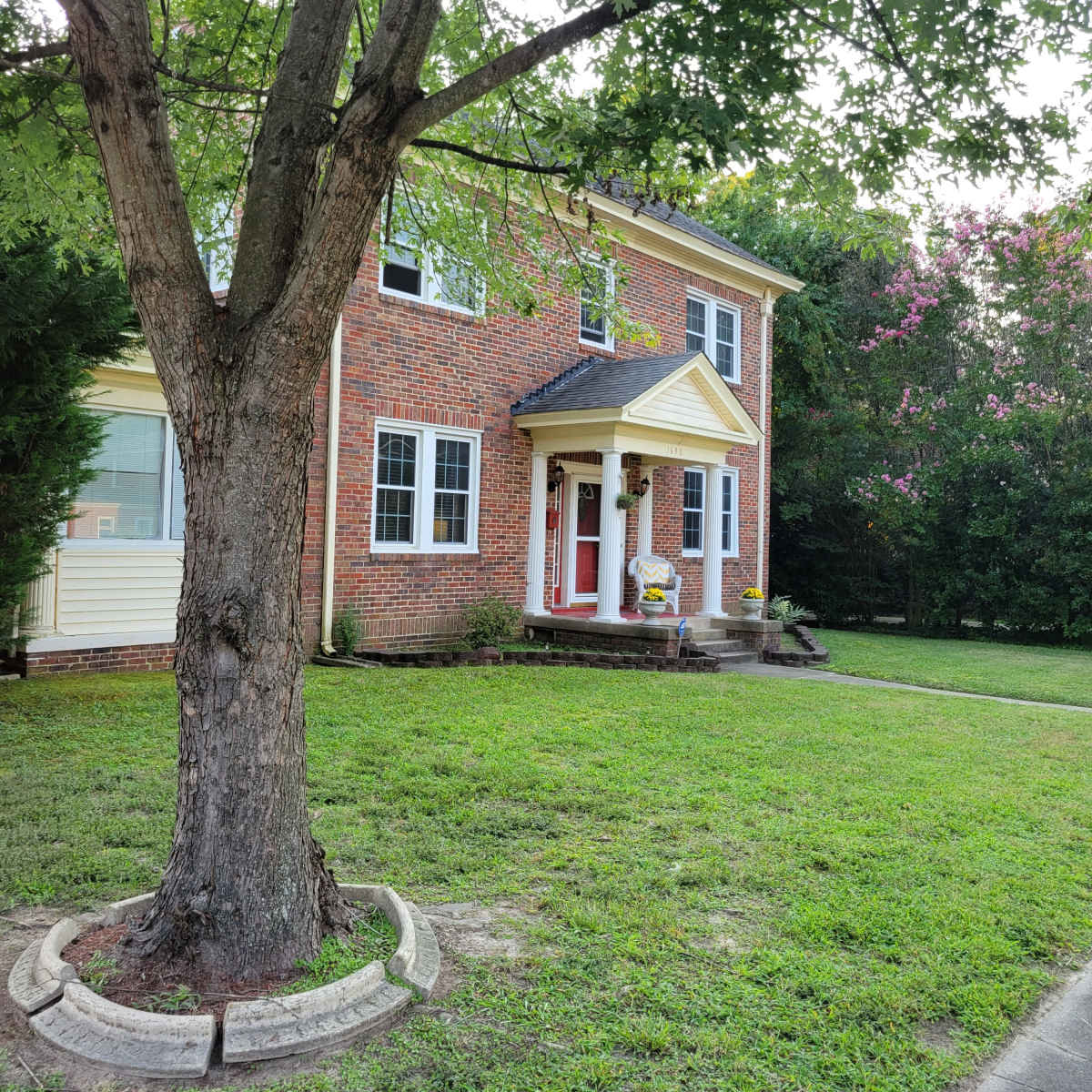 A two-story brick house with a red front door is surrounded by green grass and trees.