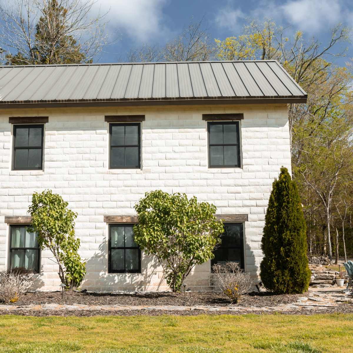 A two-story house with a metal roof and white painted block exterior is surrounded by greenery and landscaping.