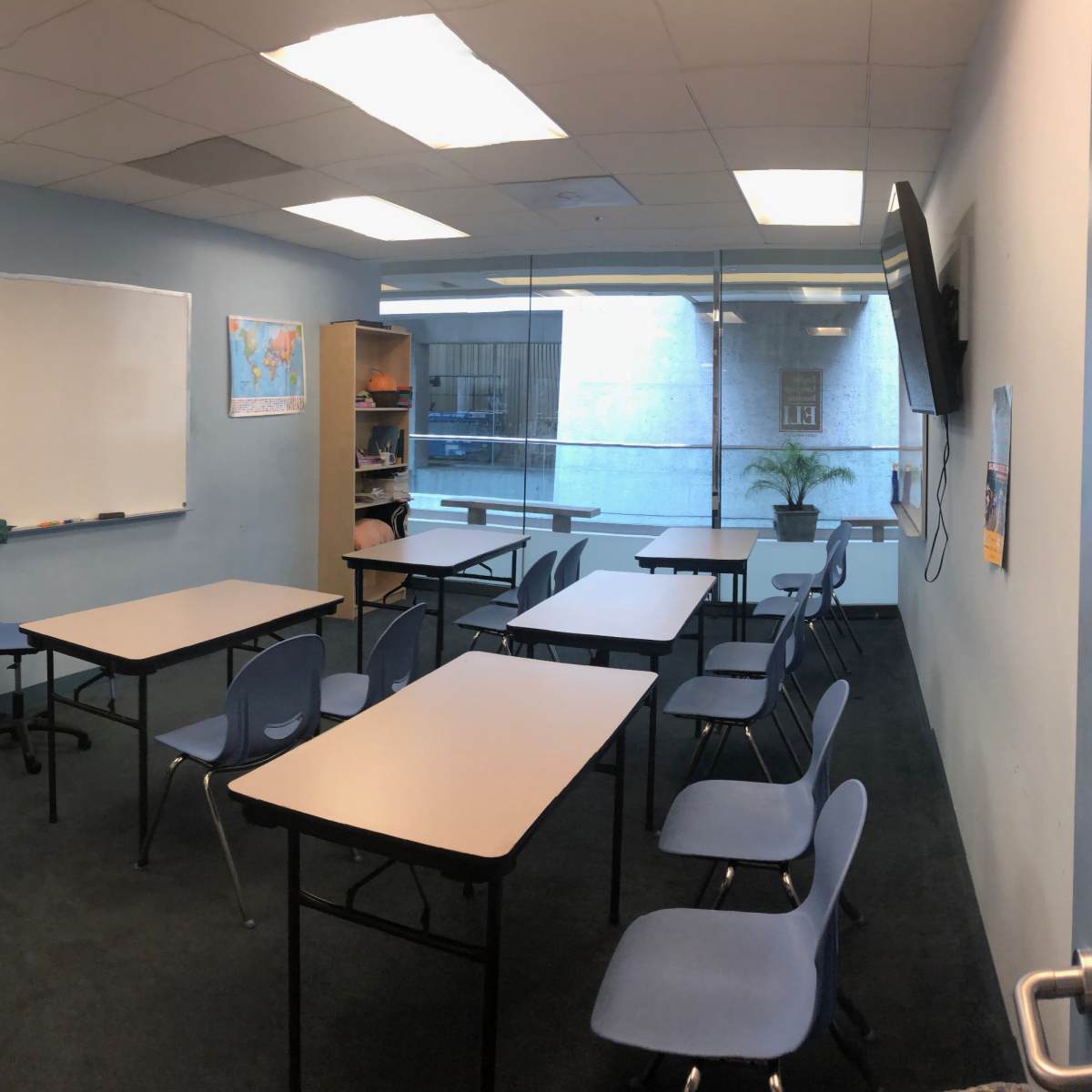 A classroom with blue walls features several tables and chairs arranged around a whiteboard and a clock on the wall.