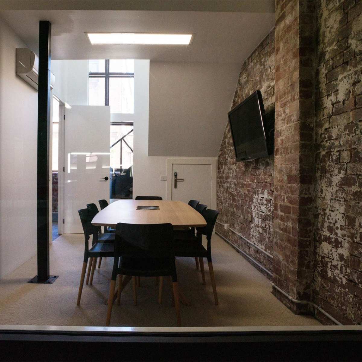 A modern conference room with a wooden table surrounded by black chairs, set against a backdrop of a brick wall and large windows.