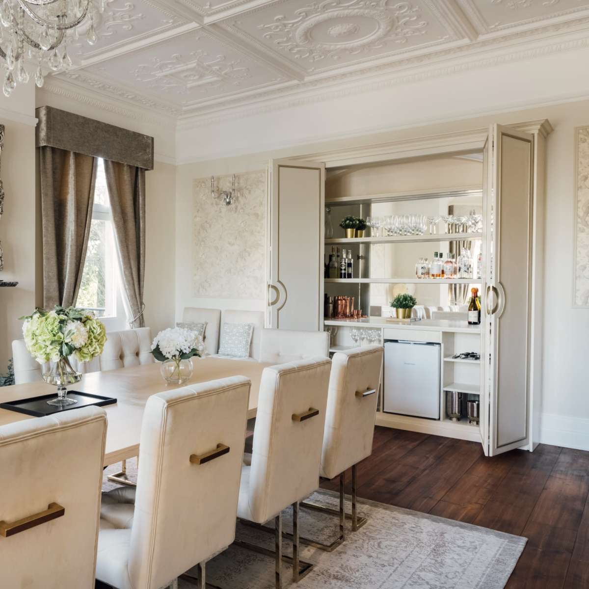 A formal dining room featuring a long wooden table surrounded by upholstered chairs, with a chandelier and a mirrored cabinet displaying glassware in the background.
