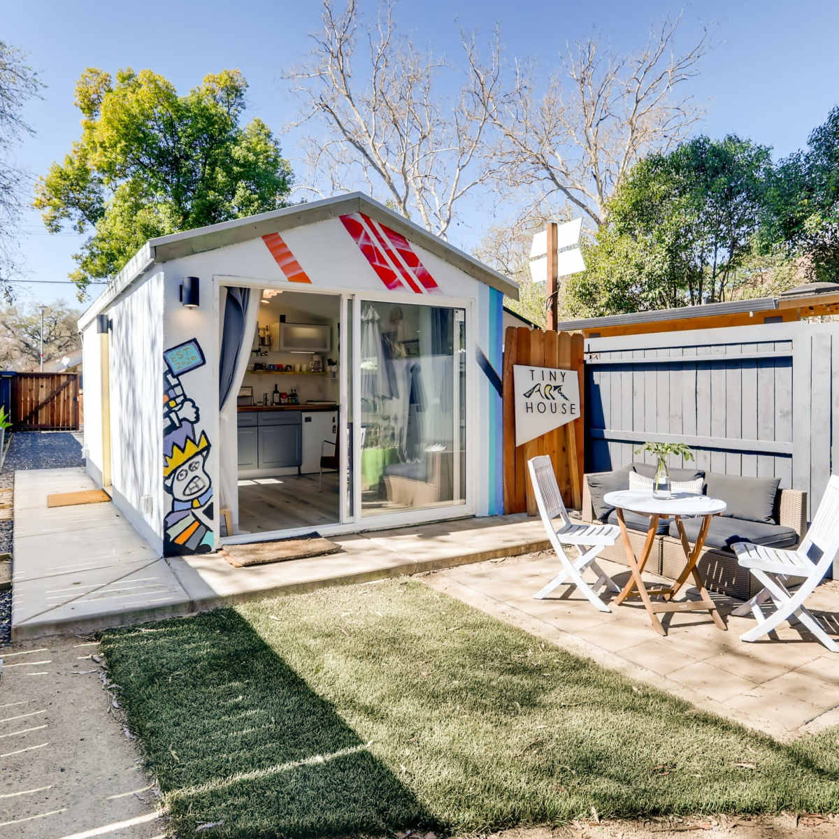 A small, modern shed-like structure with a bright mural and a patio featuring a table and chairs sits in a yard surrounded by greenery.