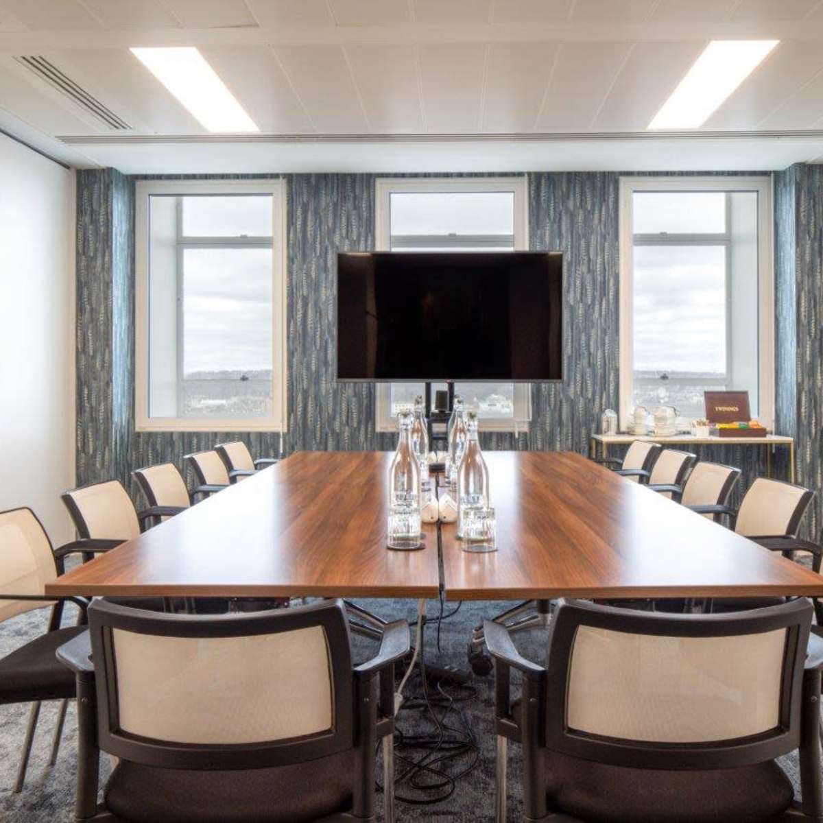 A modern conference room features a long wooden table surrounded by chairs, with a large monitor mounted on the wall and windows letting in natural light.