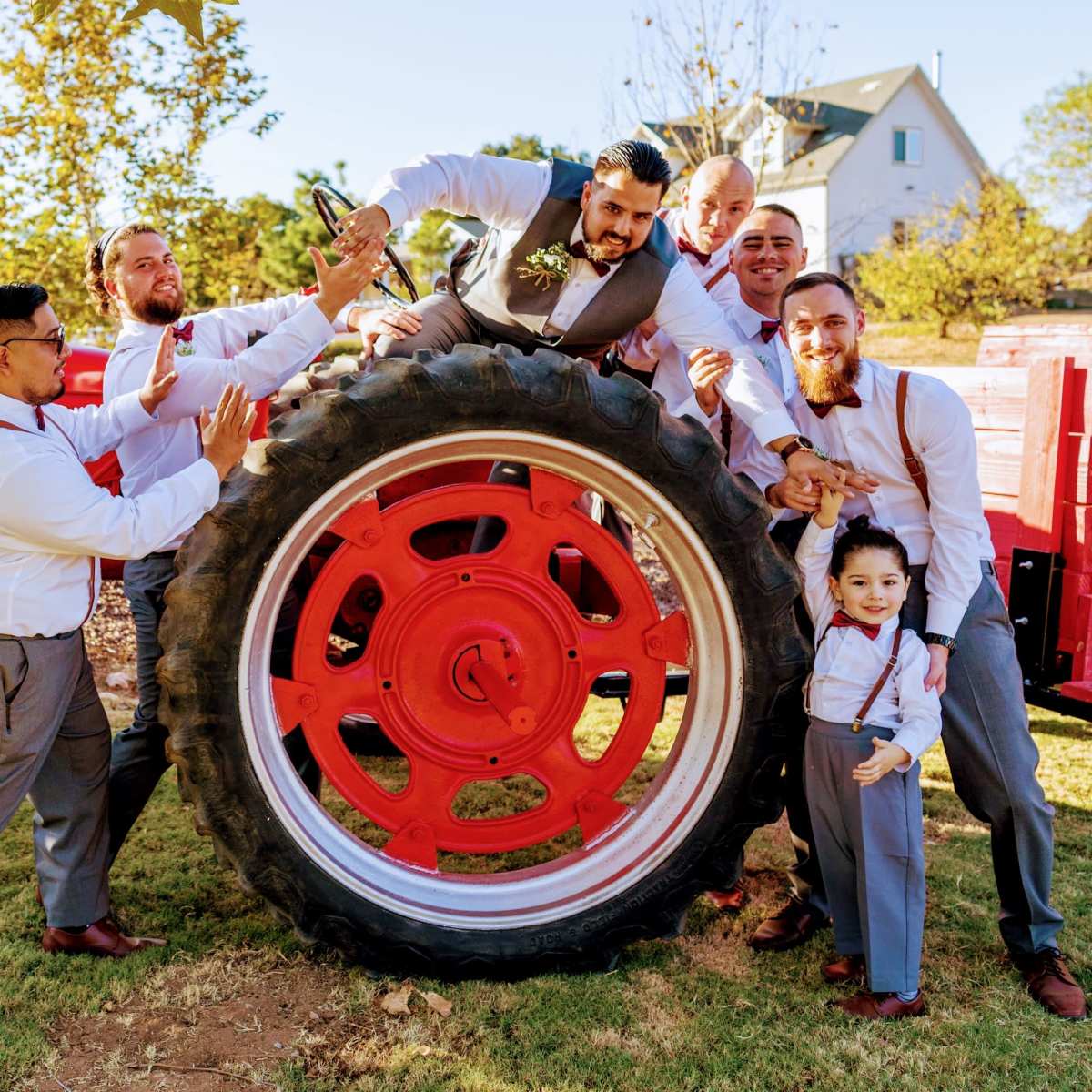 A group of men poses playfully around the large wheel of a red tractor in a grassy area.