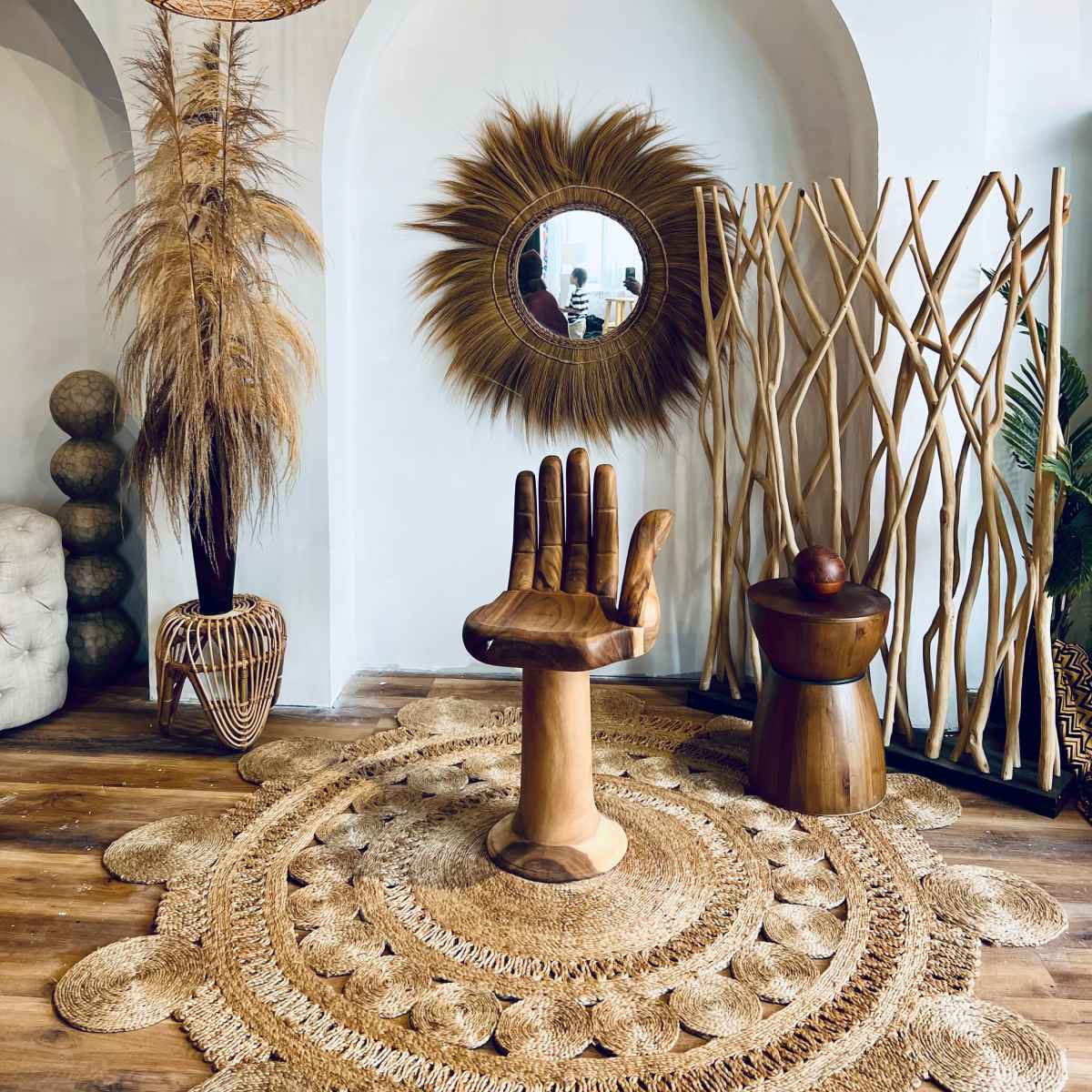 A room with a decorative wooden hand sculpture as a chair, surrounded by natural decor elements like a woven pendant light, a circular mirror, and tall bamboo elements.