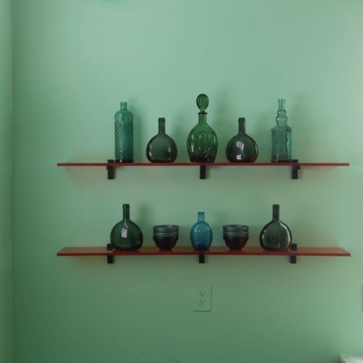 Three shelves hold various glass bottles and bowls against a light green wall with a wavy top edge.