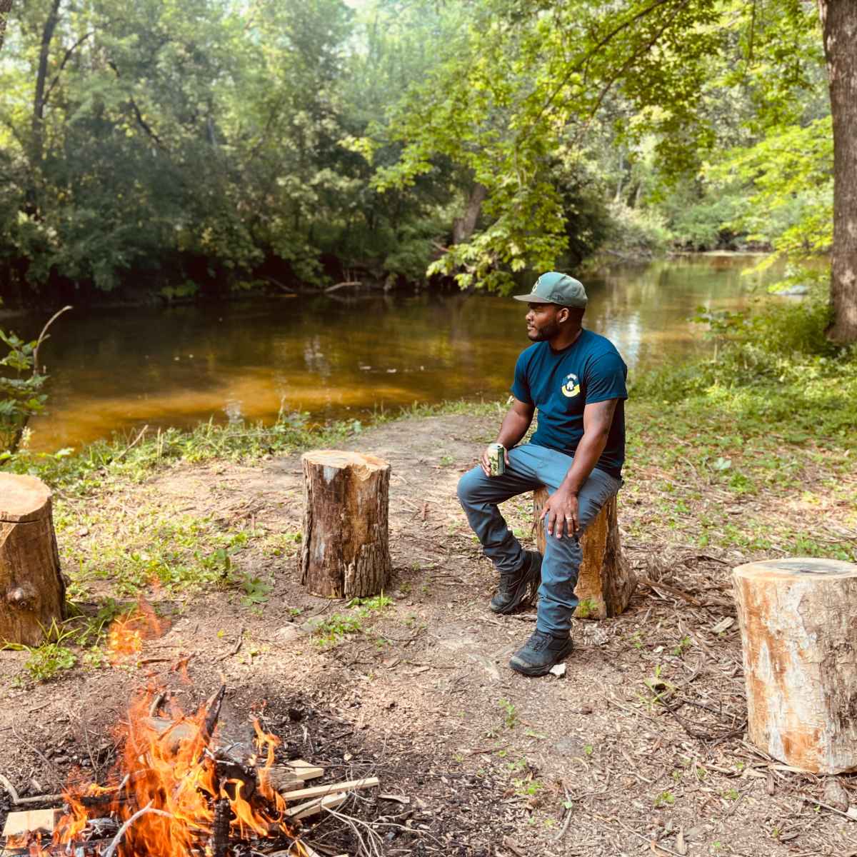 A man sits on a log beside a campfire near a calm river in a wooded area.