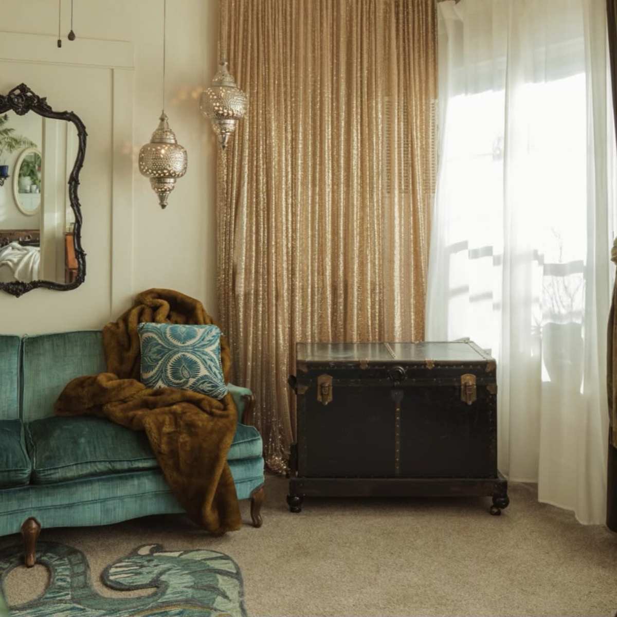 A cozy living room corner with a teal velvet sofa, a decorative throw, an antique trunk, and ornate light fixtures, all framed by golden curtains.