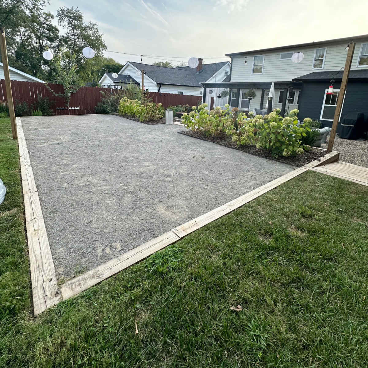A gravel area bordered by wooden planks, surrounded by flower beds and a house in the background.