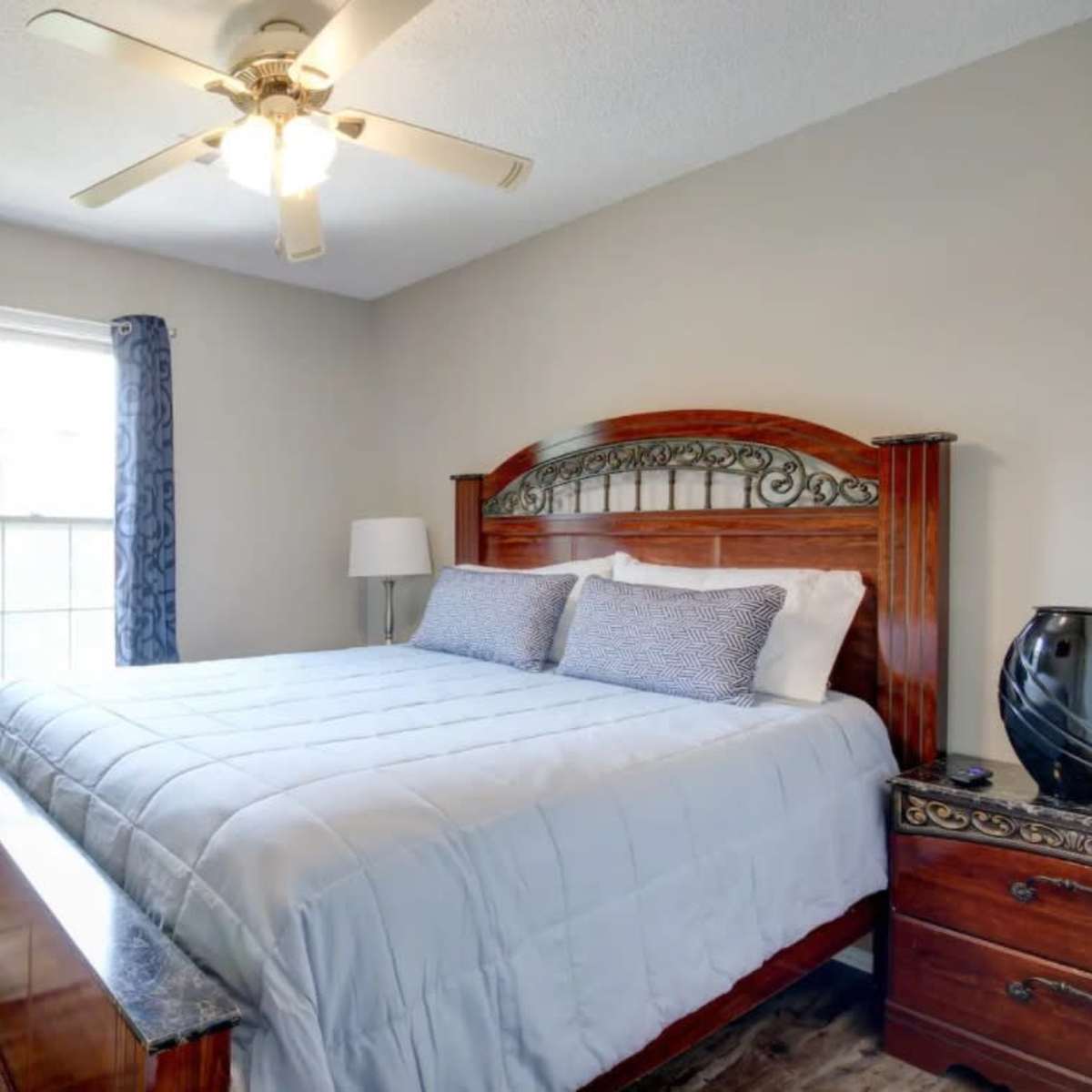 A well-lit bedroom features a wooden bed with a light blue quilt, a decorative lamp, and a window with drapes.