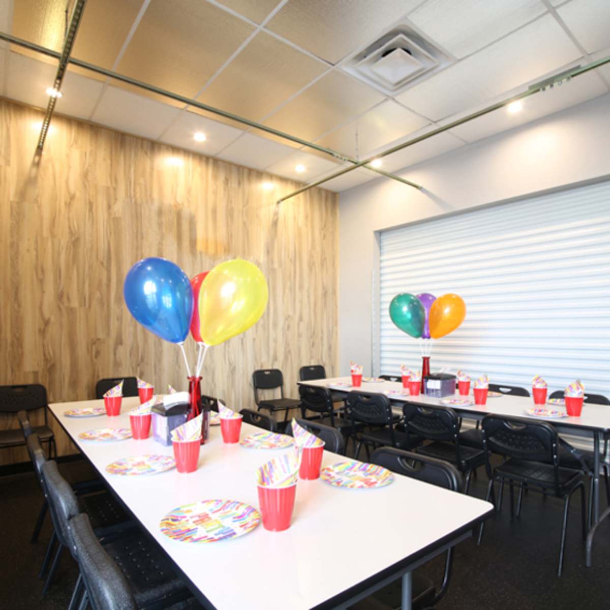 A decorated party room features a long table set with colorful plates and cups, surrounded by chairs and adorned with balloons.