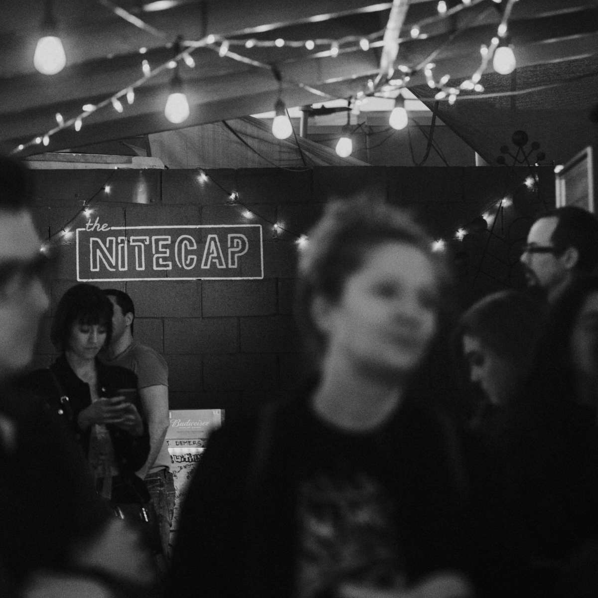 A dimly lit bar scene features a sign reading "the Nitecap," surrounded by patrons engaged in conversation under string lights.