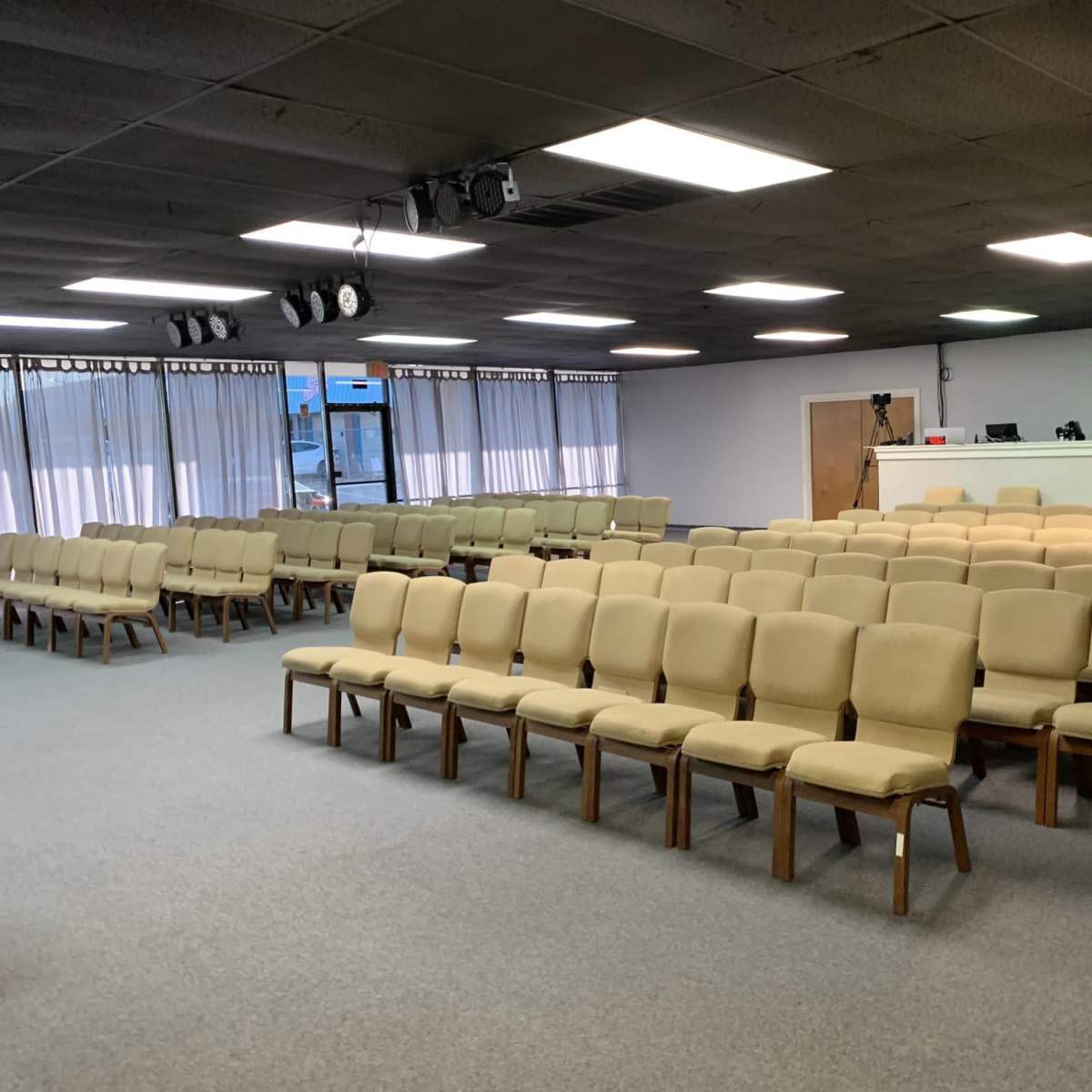 A spacious indoor area with rows of beige cushioned chairs arranged facing a blank wall and a reception desk.