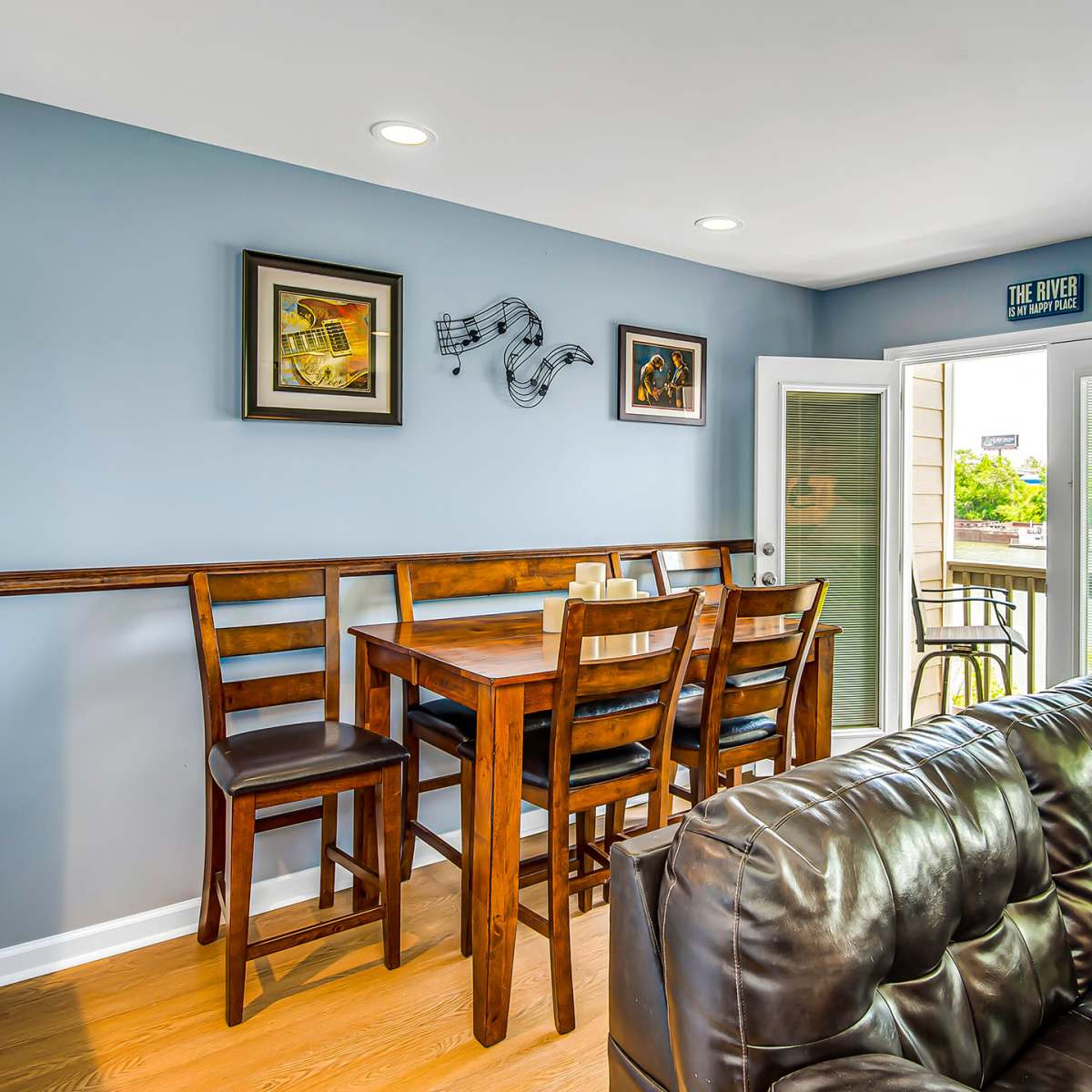 A dining area with a wooden table and chairs is positioned against a blue wall, featuring framed artwork and a door leading to an outdoor area.