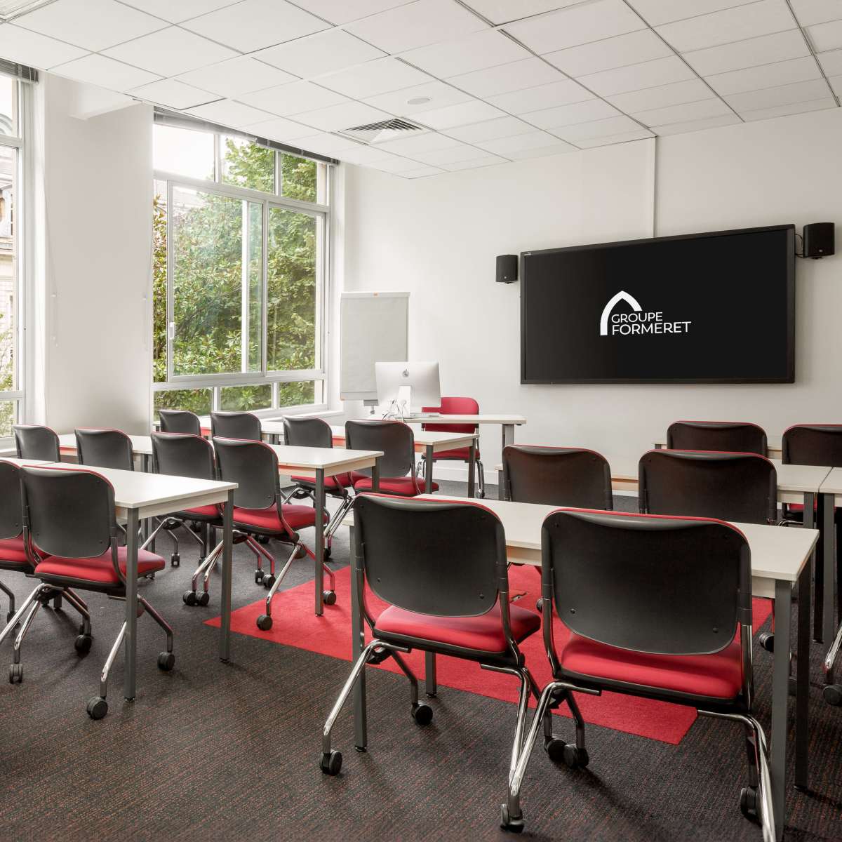 A modern classroom with rows of chairs and tables arranged facing a presentation screen, surrounded by large windows with views of greenery.