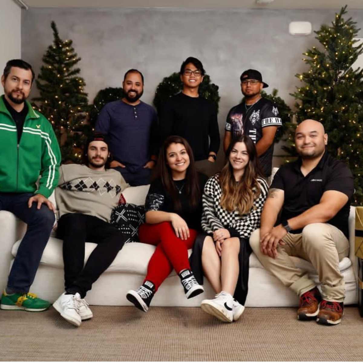 A group of eight people is posing on a couch in front of two decorated Christmas trees and gift boxes.