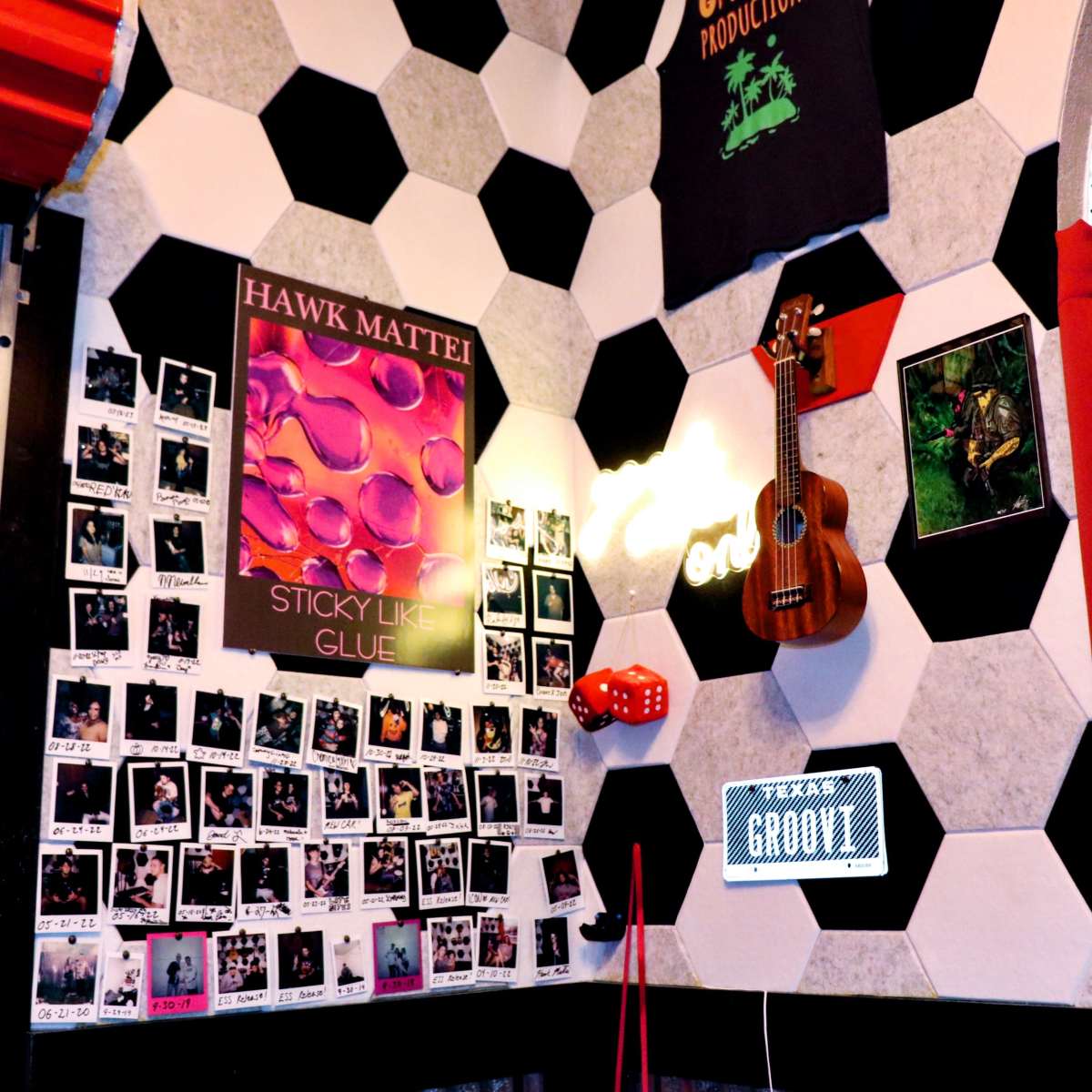 A room with soccer ball-patterned walls, a poster, a ukulele, various polaroid photos, and a light-up sign.