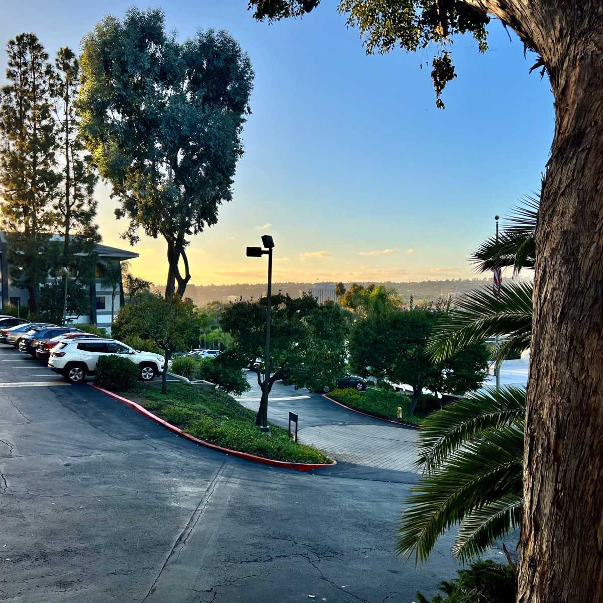 A parking lot surrounded by trees and buildings, with a view of hills in the background under a sunset sky.