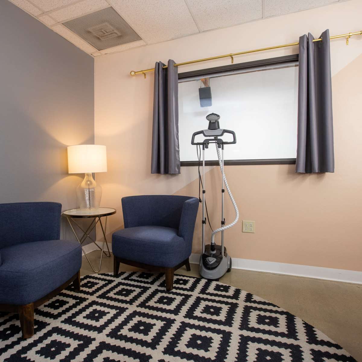 A corner of a room featuring two blue chairs, a side table with a lamp, and a garment steamer beside a window with gray curtains.