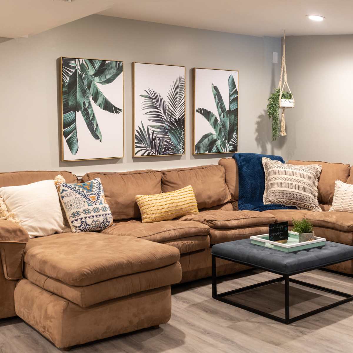A cozy living room features a large brown sectional sofa arranged around a low coffee table, complemented by wall art depicting tropical plants.