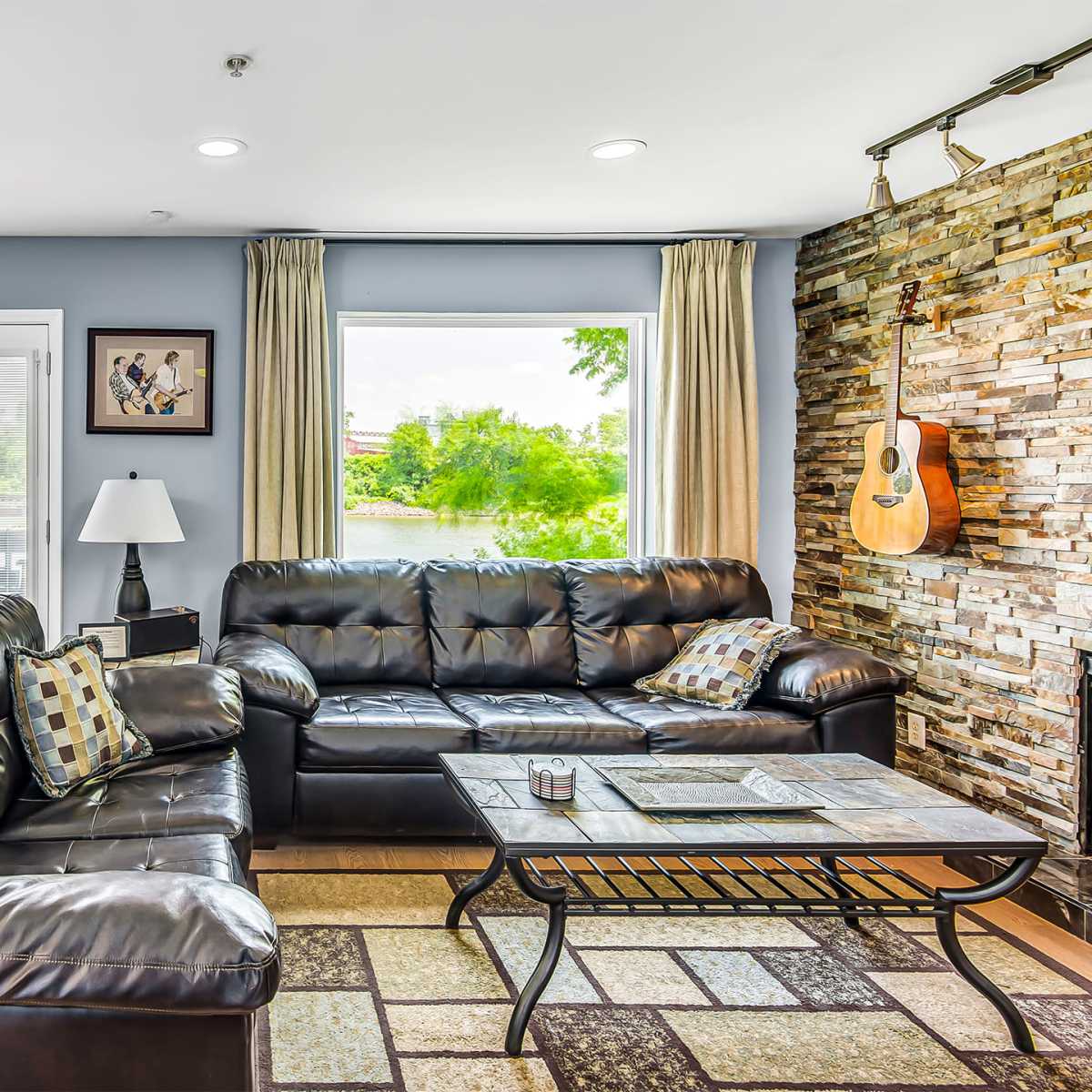 The living room features black leather sofas, a coffee table, and a stone accent wall, with a large window overlooking a scenic view.