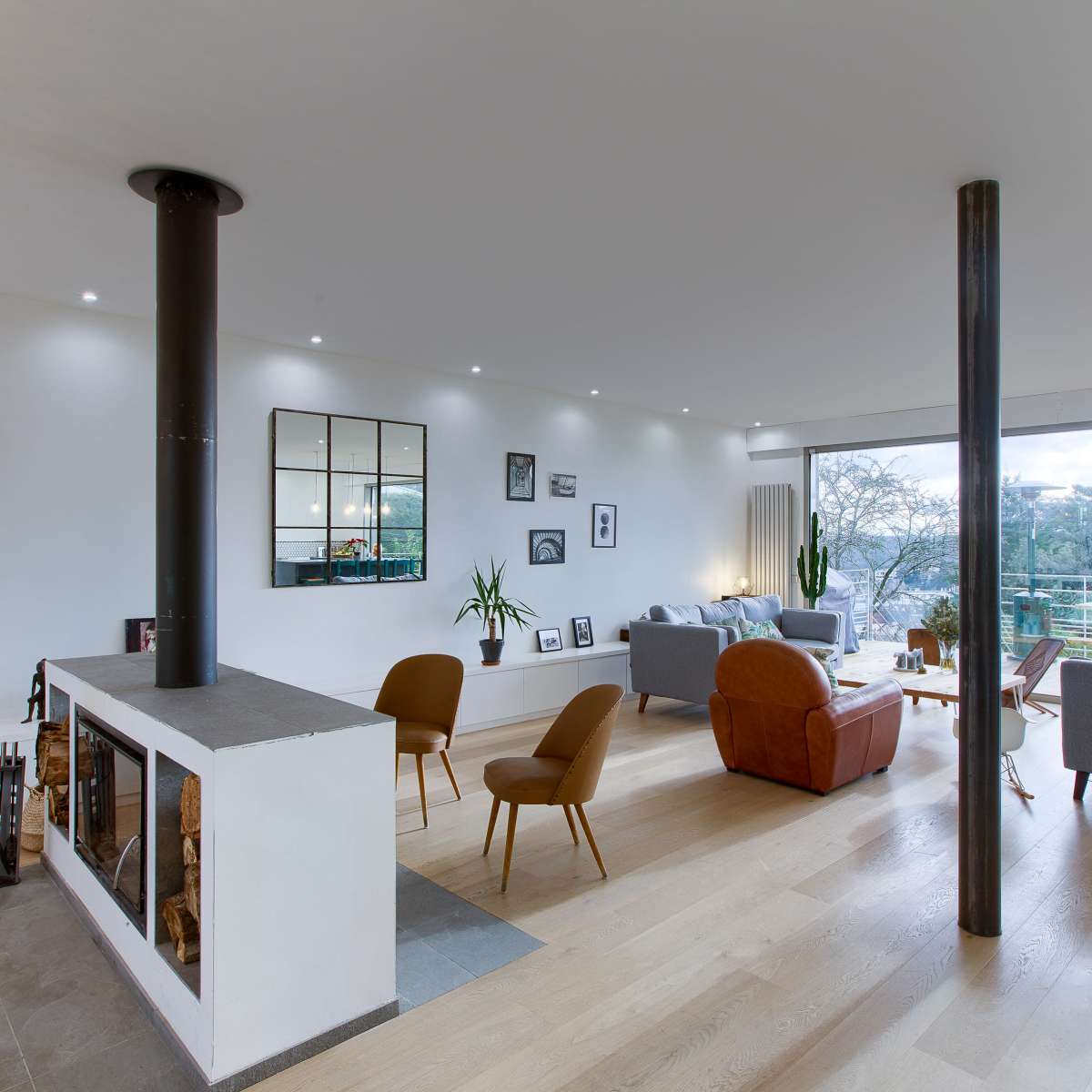 A modern living room features a central fireplace, large windows offering a view, and a mix of comfortable seating arrangements.