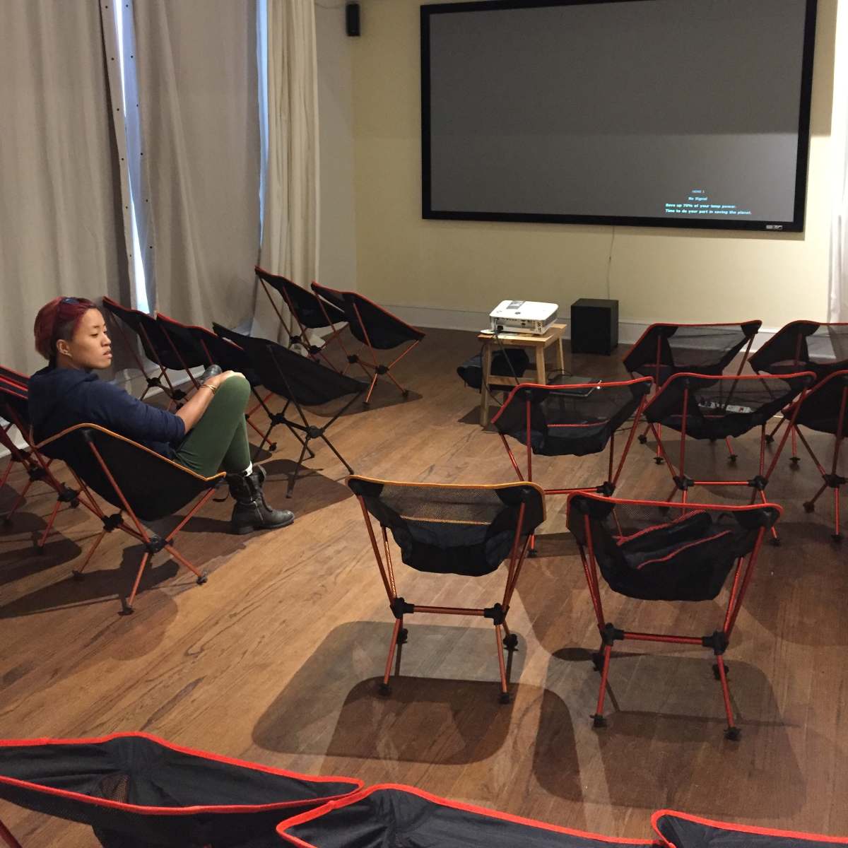 A woman sits in a nearly empty room filled with folding chairs facing a blank screen.