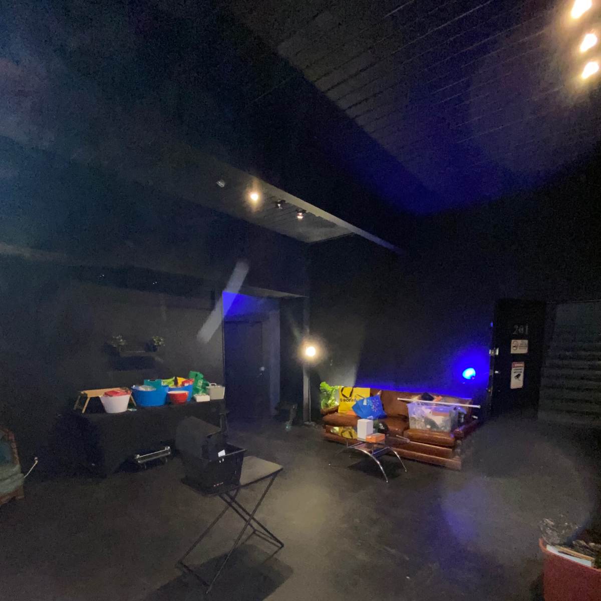 A dimly lit room with a dark floor, a chair, and various containers scattered around, along with a few light sources illuminating different areas.