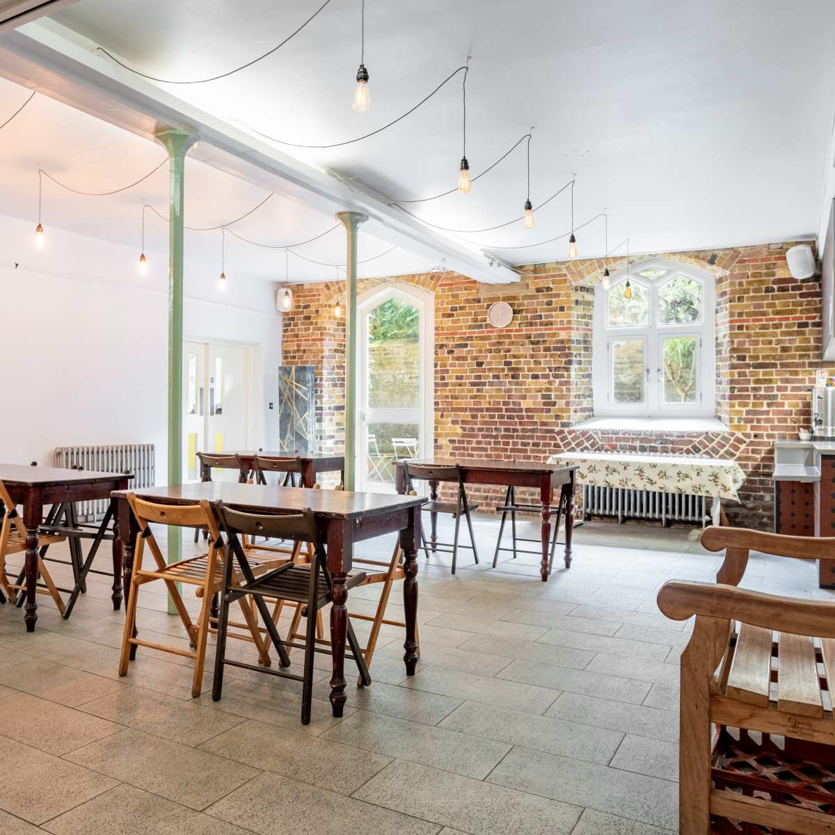 A spacious dining area with wooden tables and chairs, brick walls, and large windows, illuminated by hanging light bulbs.